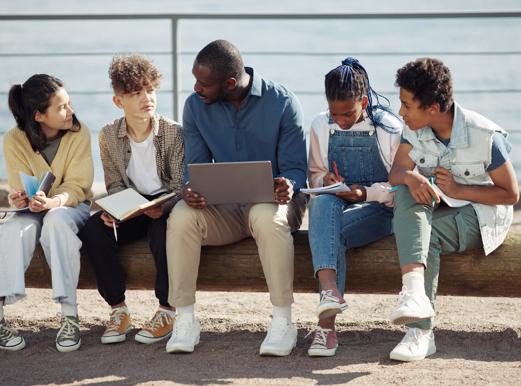 An educator collaborating with four students outdoors on a bench, reviewing coursework on a laptop while students take notes in notebooks.