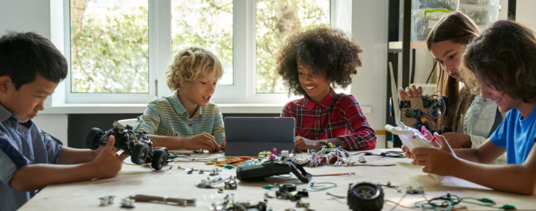 A group of five diverse children gathered around a large table in a brightly lit room, collaboratively working on small robotic cars with a tablet, remote controls, and various electronic components spread out before them.