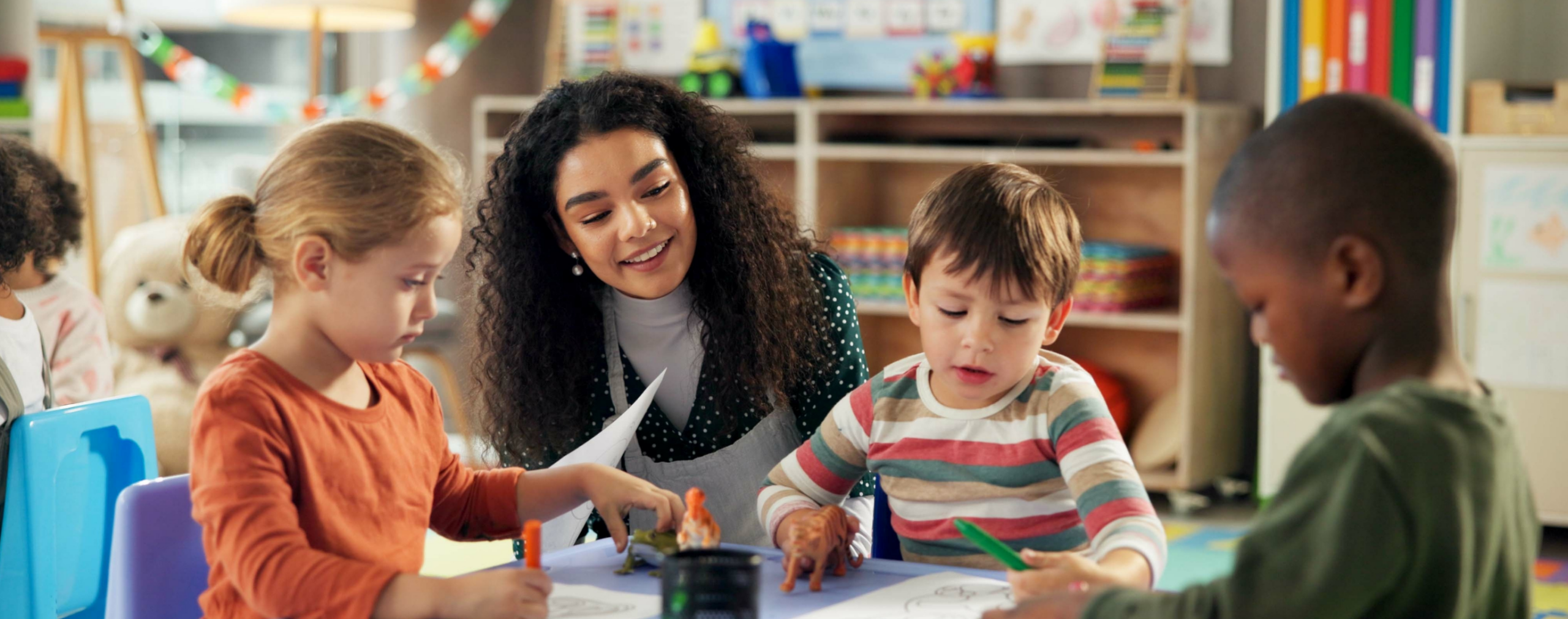 A smiling teacher sits at a small table in a bright, colorful classroom, engaging with three young children who are drawing on paper and playing with toy dinosaurs.