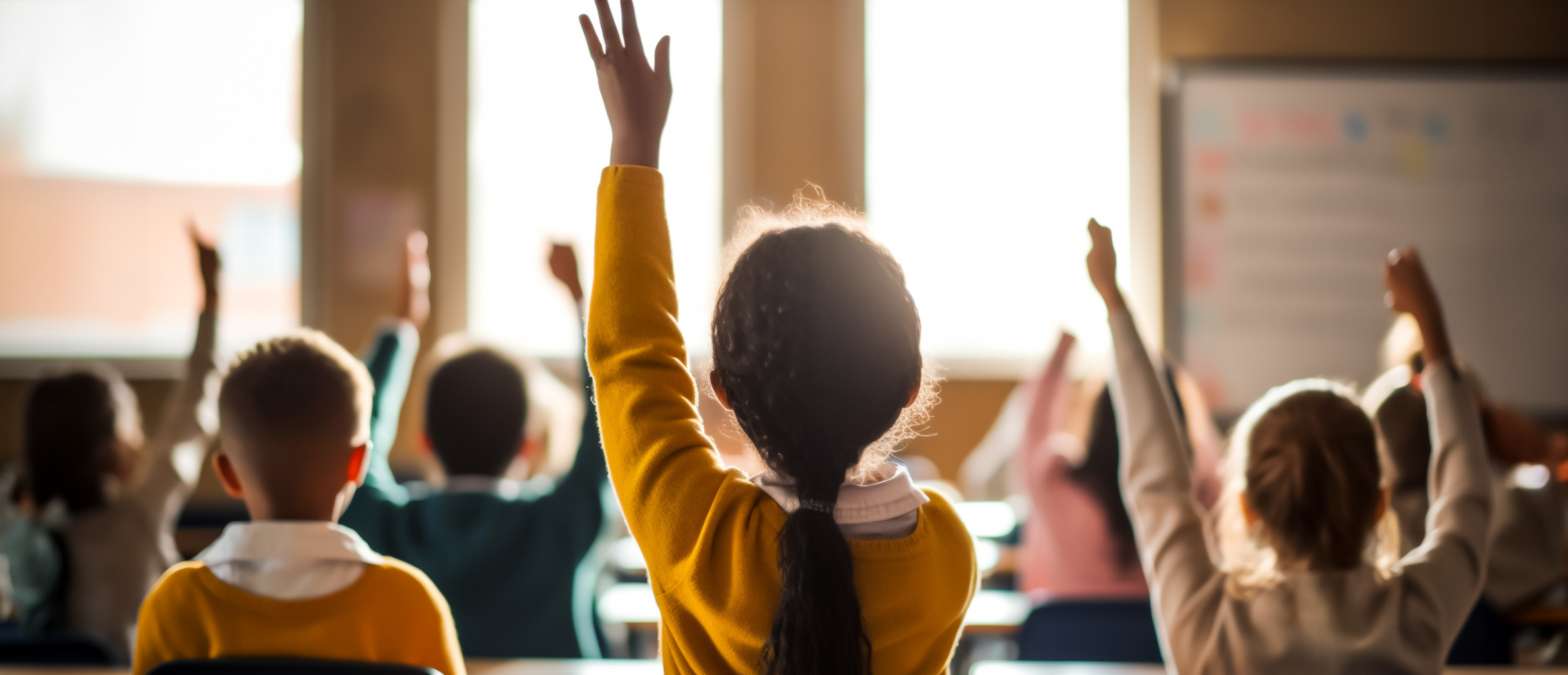 Elementary or middle school students sitting at desks with their hands raised high in a bright, sunlit classroom, demonstrating active participation and student engagement.