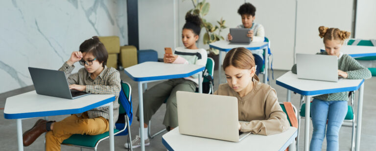 Diverse group of young children using laptops while sitting at desks in school classroom, copy space