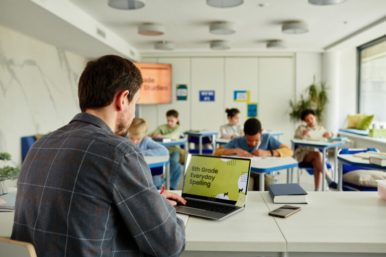 Adult white male teacher on a laptop with back turned away from point of view in a classroom with child students focusing